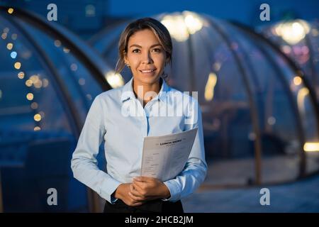 Junge hübsche Kellnerin in Uniform hält Papiermenüs, während sie vor der Kamera im großen modernen Café steht Stockfoto