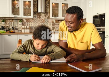 Glücklicher junger afrikanischer Mann und sein biracial Sohn mit Stift und Copybook Durchführung Hausaufgaben von Holztisch Stockfoto