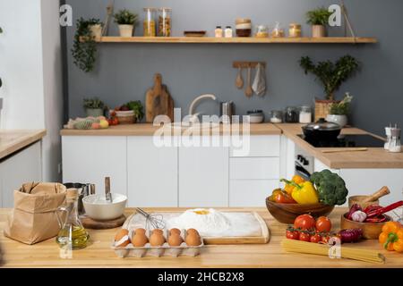 Innenraum der Küche mit Holztisch, frisches Gemüse, gesiebtes Mehl, Eier, Flasche Olivenöl und ein paar Spaghetti Stockfoto