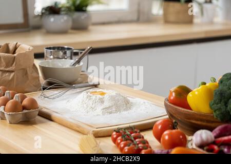 Haufen gesiebenem Mehl mit rohem Ei auf der Oberseite und anderen Zutaten für hausgemachtes Gebäck auf dem Küchentisch Stockfoto