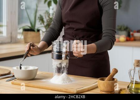 Hände einer jungen Frau in der Schürze, die Mehl für hausgemachtes Gebäck über Holzbrett sieben, während sie am Küchentisch steht Stockfoto