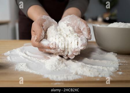 Hände einer jungen Frau, die einen Haufen gesiebenem Mehl auf einem Holzbrett hält, während sie in der Küche hausgemachtes Gebäck zubereitet Stockfoto