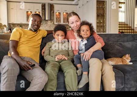 Junge, liebevolle Familie, die auf der Couch sitzt und Filme auf dem fernseher ansieht, während sie zu Hause bleibt und sich ausruhen kann Stockfoto