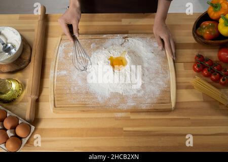 Junge Frau, die Schneebesen über einen Haufen gesiebenem Mehl mit rohem Eigelb auf der Oberseite hält, während sie den Teig für hausgemachtes Gebäck zubereitet Stockfoto