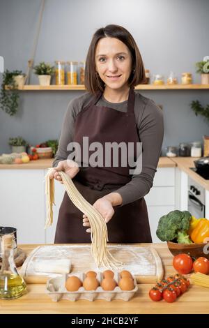 Glückliche junge kaukasische Frau in Schürze Vorbereitung Spaghetti, während sie über Holzbrett halten Stockfoto