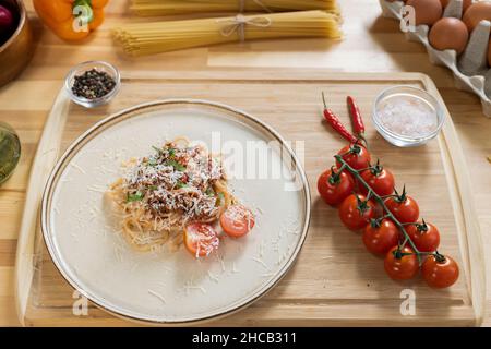 Stapel von Carbonara-Nudeln, die mit geriebenem Käse auf dem Teller, umgeben von frischen Tomaten, Salz und Pfeffer in kleinen Schüsseln, bestreut werden Stockfoto