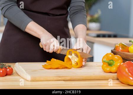 Hände junger Frauen hacken frische gelbe Paprika auf Holzbrett, während sie Gemüseeintopf zubereiten Stockfoto