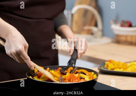 Hände einer jungen Frau, die Gemüseeintopf mit Holzlöffel mischt, während sie die Bratpfanne am Griff hält Stockfoto