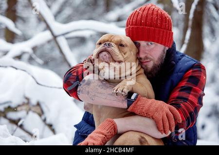 Glücklicher Mann, der in einem verschneiten Wald einen schönen Hund in den Händen hält. Lächelnder Junge umarmt entzückenden Welpen im Winterholz. Tierliebhaber. Hund - Mensch s Freund Konzept Stockfoto