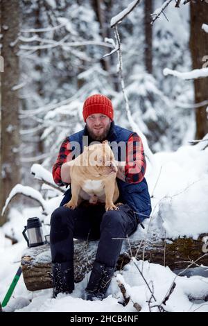 Glücklicher Mann, der in einem verschneiten Wald einen schönen Hund in den Händen hält. Lächelnder Junge umarmt entzückenden Welpen im Winterholz. Tierliebhaber. Hund - Mensch s Freund Konzept Stockfoto