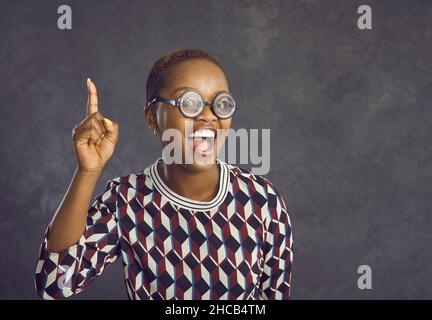 Fröhliche Frau in lustigen Gläsern hebt ihren Zeigefinger, um anzuzeigen, dass sie eine Idee oder einen Gedanken hat. Stockfoto