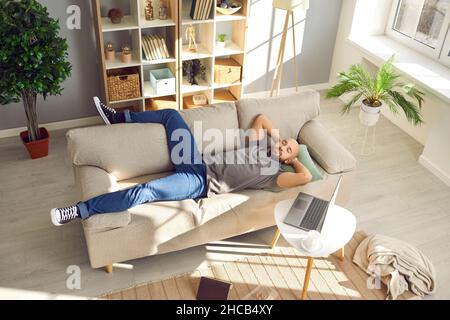 Mann, der sich von der Arbeit auf dem Laptop ausbrach und auf der bequemen Couch im Wohnzimmer lappte Stockfoto