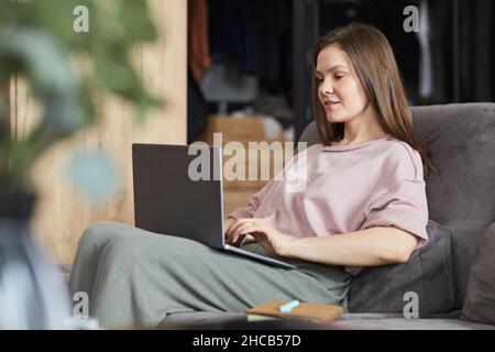 Junge Frau, die auf dem Sofa sitzt und auf dem Laptop tippt, arbeitet zu Hause online Stockfoto