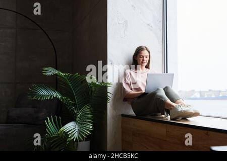 Junge Frau, die auf der Fensterbank sitzt und online mit ihrem Laptop im Zimmer arbeitet Stockfoto