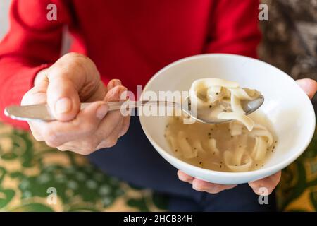 Oben Ansicht der Schüssel mit Hühnernudelsuppe in der Hand der alten Frau (konzentrieren Sie sich auf Nudeln in Esslöffel) Stockfoto