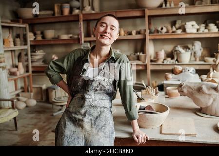 Porträt einer jungen glücklichen Frau im Schürze, die während der Arbeit in einer Keramikwerkstatt die Kamera anlächelt Stockfoto
