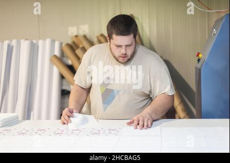 Der Mitarbeiter des Logistikdruckers steuert und ordnet das gedruckte Material auf der Produktionslinie in der digitalen Druckerei. Industriearbeiten Stockfoto