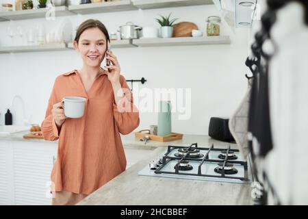 Junge Frau, die morgens in der Küche Kaffee trinkt und ein Gespräch mit dem Handy führt Stockfoto