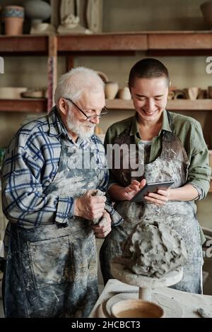 Junge Frau, die einen Tablet-pc benutzt und dem älteren Mann etwas zeigt, während er während ihrer Teamarbeit in der Töpferwerkstatt Ton in den Händen modelliert Stockfoto