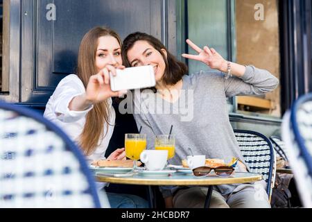 Junge Frau, die Selfie mit einem Freund gemacht hat und ein Friedensschild im Café zeigt Stockfoto