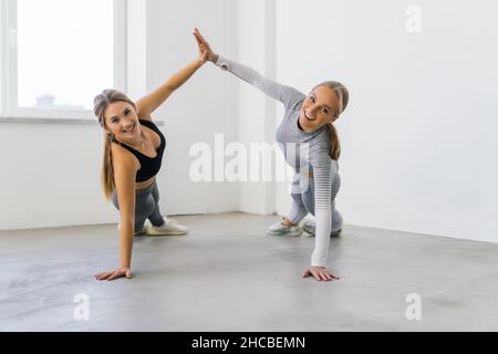 Junge Frauen üben zusammen Yoga, geben hohe fünf Hände und stehen in der Seitenplanke Pose auf Gummimatten im Fitnessstudio Stockfoto