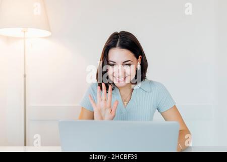 Lächelnde junge Geschäftsfrau, die im Heimbüro über einen Laptop mit Videoanruf winkt Stockfoto