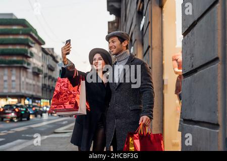 Lächelndes junges Paar mit Einkaufstaschen, das Selfie mit dem Smartphone in der Nähe des Geschäfts nimmt Stockfoto
