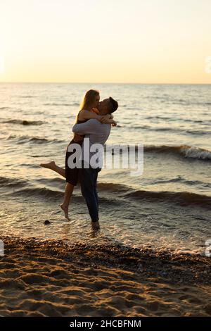 Romantischer Mann hebt und küsst Frau am Strand bei Sonnenuntergang Stockfoto