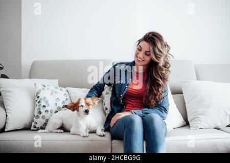 Lächelnde Frau mit langem Haar streichelnder Hund auf dem Sofa Stockfoto