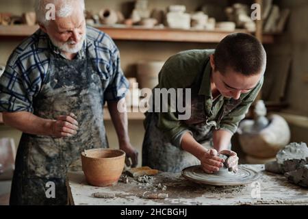 Eine Künstlerin formt und modelliert eine Töpfervase aus rohem Ton zusammen mit einem älteren Mann, der ihr im Atelier assistierte Stockfoto