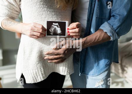 Lächelnder Mann mit einer Schwangeren, die im Wohnzimmer zu Hause Ultraschalluntersuchungen vorführt Stockfoto