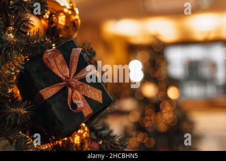 Samt-Neujahrsschachtel mit goldenem Glitzerband, das mit goldenen Kugeln am festlichen Weihnachtsbaum hängt. Magische Girlande Lichter und Winter holida Stockfoto