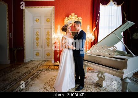Der Bräutigam und die Braut umarmen und küssen sich in der Nähe des Klaviers im luxuriösen Interieur Stockfoto