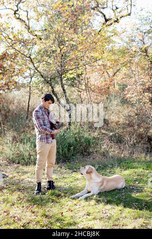 Junger Mann mit Feuerholz beim Blick auf Labrador Retriever im Herbstwald Stockfoto