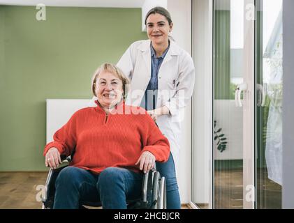 Lächelnde Krankenschwester, die zu Hause im Rollstuhl hinter einer Frau steht Stockfoto