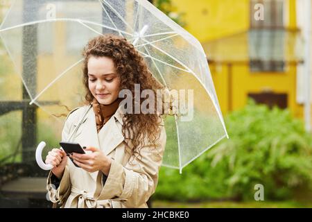 Horizontales mittleres Porträt einer jungen Frau im Trenchcoat, die an regnerischen Tagen im Freien unter einem Regenschirm steht und im Internet auf dem Smartphone surft Stockfoto
