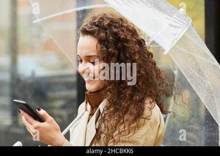 Horizontales, mittleres Nahaufnahme-Porträt einer kaukasischen Frau mit lockigen Haaren, die am regnerischen Tag unter einem Regenschirm im Freien steht und das Internet auf dem Smartphone nutzt Stockfoto