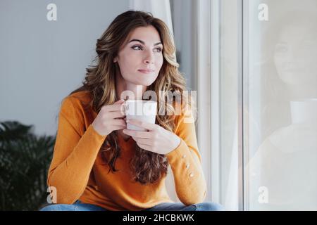 Junge Frau mit Tasse Kaffee durch die Fenster zu Hause Stockfoto