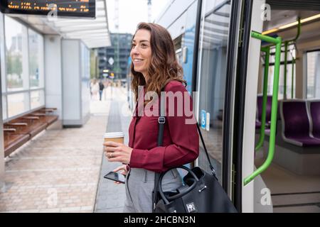 Lächelnde Geschäftsfrau mit Einweg-Becher und Mobiltelefon, die am Bahnhof aus dem Zug aussteigt Stockfoto