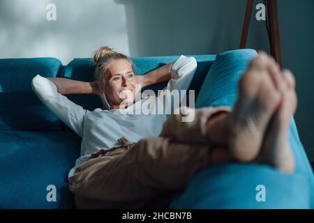 Lächelnde Frau mit Händen hinter dem Kopf auf dem Sofa Stockfoto