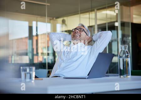 Glücklicher Geschäftsmann, der mit den Händen hinter dem Kopf im Büro sitzt Stockfoto