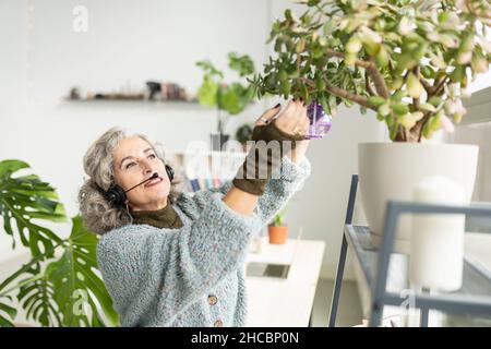 Geschäftsfrau mit Kopfhörern, die zu Hause Wasser auf die Pflanzen sprüht Stockfoto