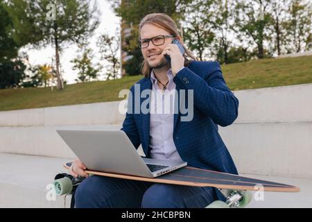 Geschäftsmann mit Skateboard und Laptop, der auf dem Mobiltelefon über die Stufen spricht Stockfoto