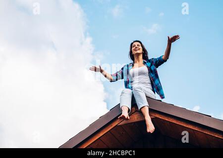 Frau mit ausgestreckten Armen sitzt auf dem Dach des Hauses Stockfoto