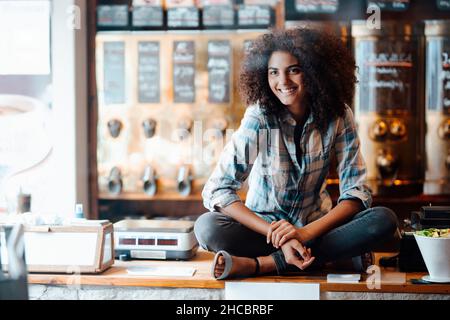 Geschäftsfrau sitzt mit gekreuzten Beinen auf der Theke bei der Kaffeerösterei Stockfoto
