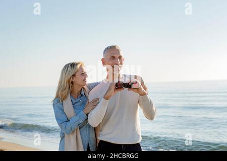 Lächelnde Frau mit Arm um den Mann, der ein Fernglas am Strand hält Stockfoto