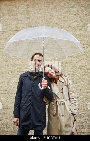 Vertikale Aufnahme eines glücklichen Paares, das im Freien unter einem Regenschirm an einer beigen Gebäudewand steht und die Kamera anschaut Stockfoto