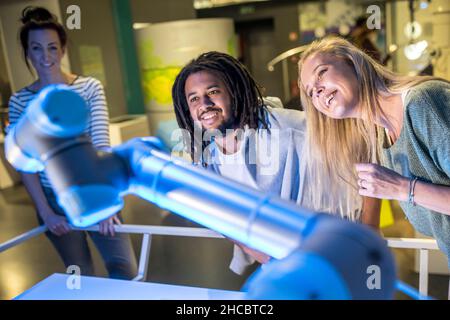 Lächelnde junge Tech-Entwickler mit Kollegen, die in der Werkstatt auf den Roboterarm schauen Stockfoto