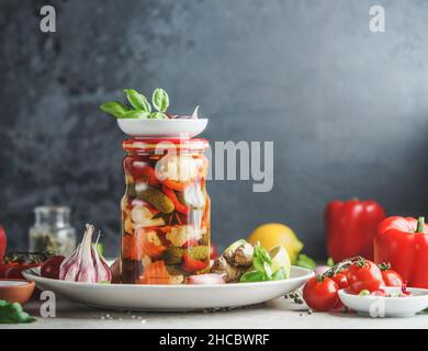 Hausgemachtes Gemüse im Glas auf weißem Teller am Tisch mit Küchenutensilien und Zutaten, Kräuter und Gemüse an schwarzer Wand Backgroun Stockfoto