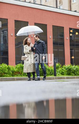 Vertikale Langaufnahme des kaukasischen Mannes und der Frau, die am regnerischen Frühlingstag unter einem Regenschirm auf der Straße spazieren Stockfoto